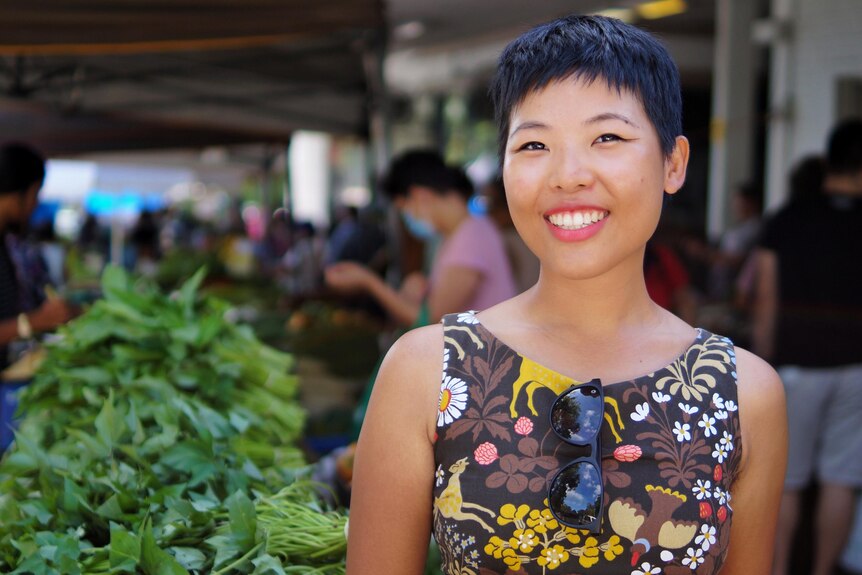 Tisha stands in front of a market stall table piled high with leafy green vegetables while wearing a brown dress and smiling.