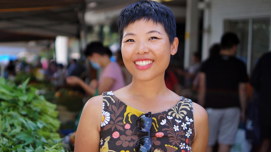 Tisha stands in front of a market stall table piled high with leafy green vegetables while wearing a brown dress and smiling.