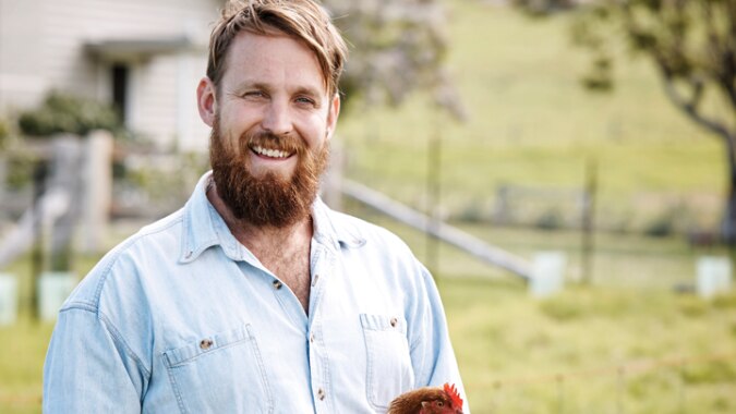 A bearded man stands on a farm holding a red hen.