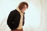 Lewis Capaldi looks at his feet in a cream hoodie and black jacket with a billowing off-white backdrop.