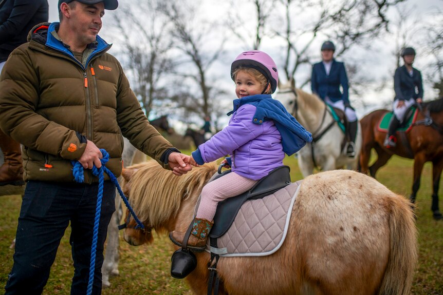 Holding onto her dad's index finger, a girl in a purple jacket and pink helmet smiles while riding a cream-coloured pony