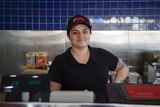 A young woman with dark hair wearing a cap that says the pantry stares down the camera from behind a takeaway counter, smiling.