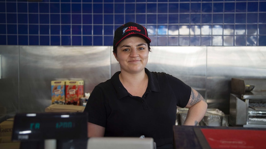 A young woman with dark hair wearing a cap that says the pantry stares down the camera from behind a takeaway counter, smiling.