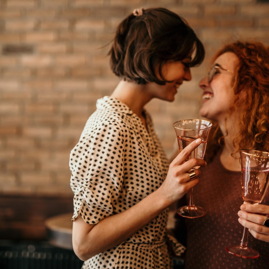 Two women at a bar holding champagne flutes and looking at each other smiling
