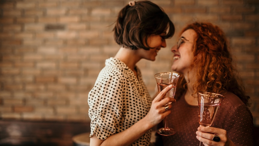 Two women at a bar holding champagne flutes and looking at each other smiling