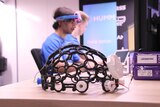 A 3D print of a prototype device designed to sit on a human head sits on a table with two men sat at computers behind it.