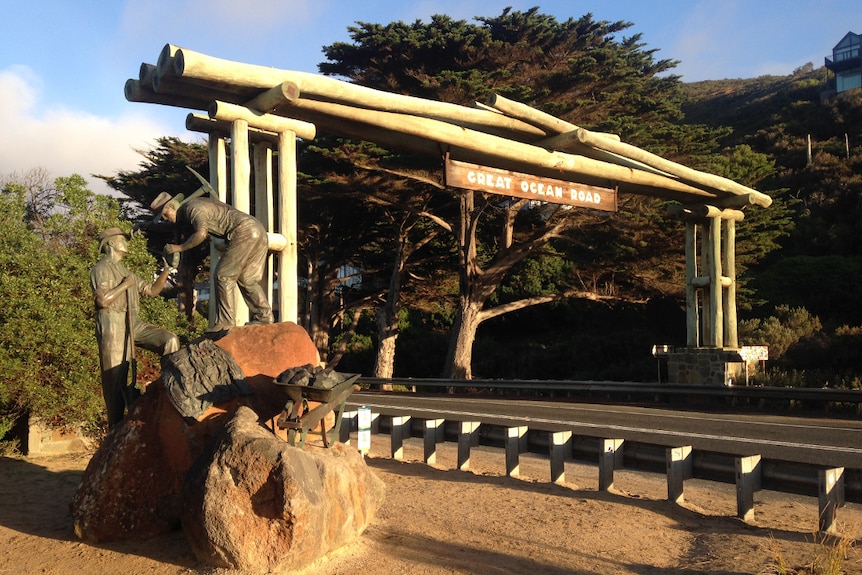 A timber arch built over the road with a sign on it saying Great Ocean Road. Beside a bronze statue of two men with tools.