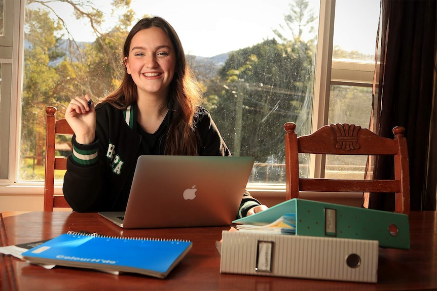 Ella Perusco sits at a dining table with an open laptop and folders, holding a pen in her school uniform.