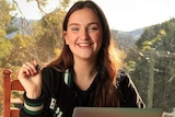 Ella Perusco sits at a dining table with an open laptop and folders, holding a pen in her school uniform.