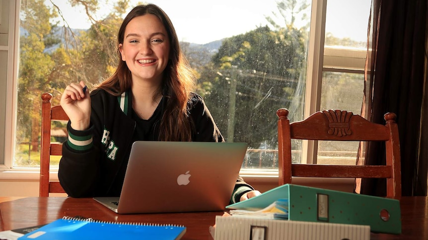 Ella Perusco sits at a dining table with an open laptop and folders, holding a pen in her school uniform.