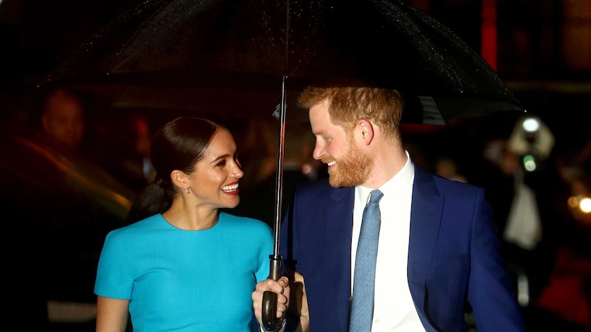 Meghan Markle wearing a blue dress smiles as she stares at Prince Harry in a dark blue suit holding an umbrella.