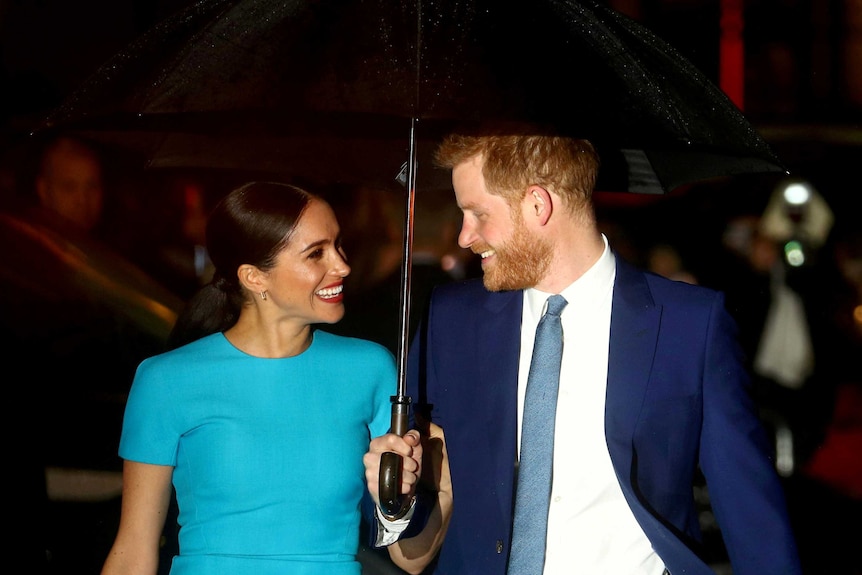 Meghan Markle in a blue dress smiles as she watches Prince Harry in a dark blue suit holding an umbrella.