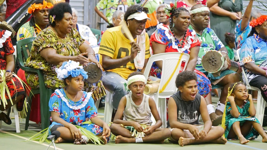 A group of adults and children sit around singing.