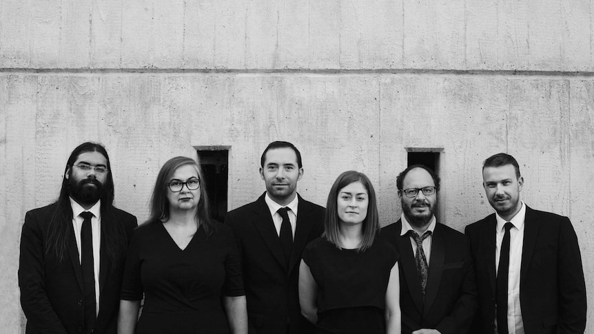 A black and white portrait of six members of "Decibel" standing in front of a concrete wall