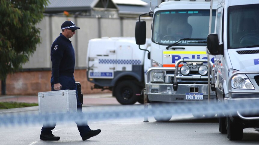 A police officer walks across a suburban road to a police van, carrying a silver trunk and a black case.