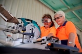 Two elderly women wearing orange high vis shirts learning how to use drop saw.