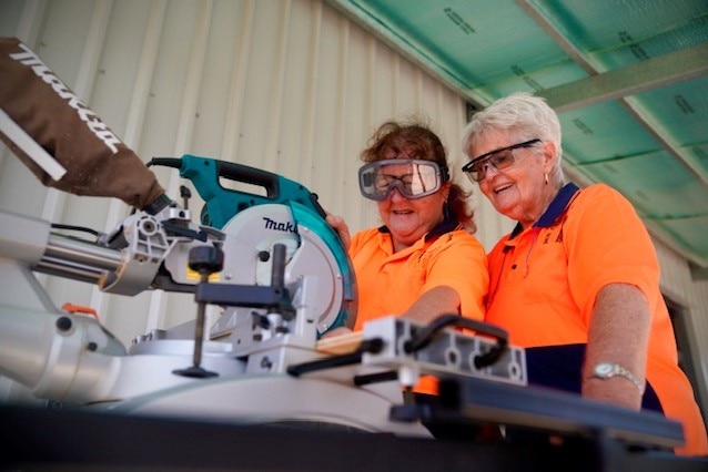 Two elderly women wearing orange high vis shirts learning how to use drop saw.