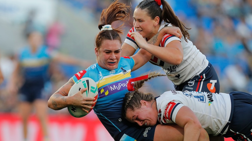 An NRLW player in blue falling backwards in a tackle with the ball, as two opponents grapple her