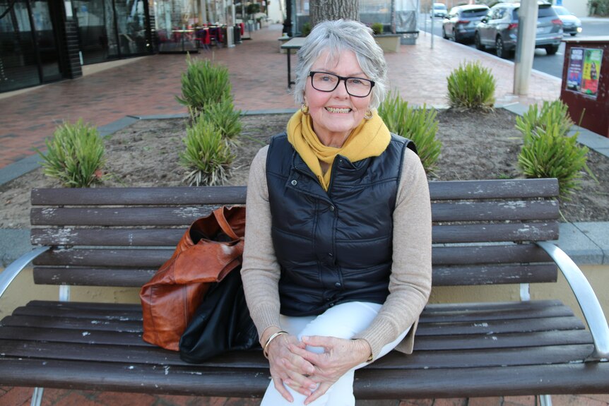 A woman sits on a bench, smiling, wearing a yellow scarf.