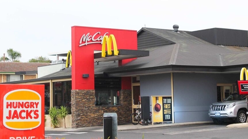 A silver car waiting in the drive-through of fast food business, McDonalds with a Hungry Jacks sign to the left of the image