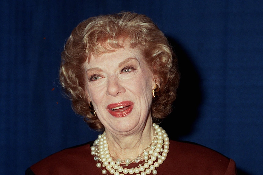 An elderly woman with blond hair, wearing pearls and smiling