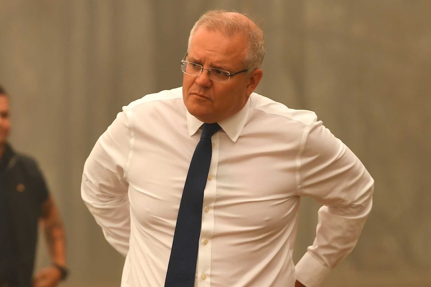 Scott Morrison stands in front of fire-ravaged bushland with a serious expression on his face.