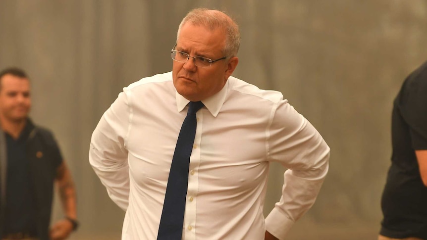 Scott Morrison stands in front of fire-ravaged bushland with a serious expression on his face.