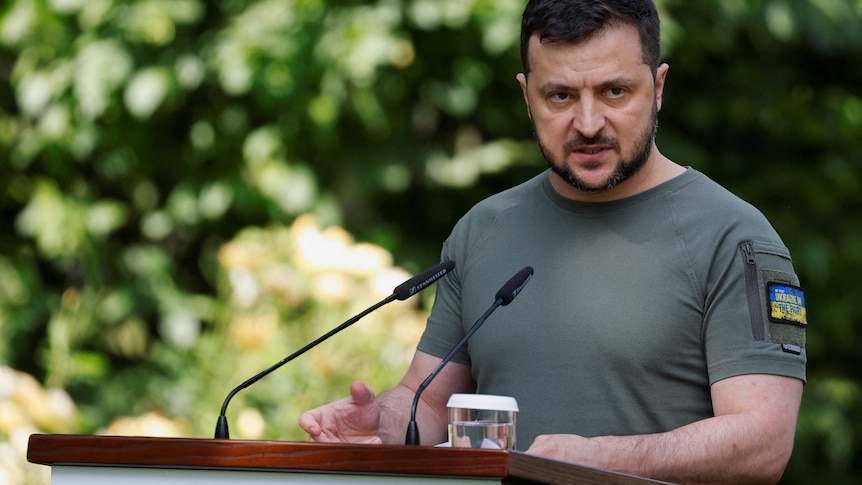 A man in a grey shirt with Ukraine flag patches on the sleeve speaks at podium. 