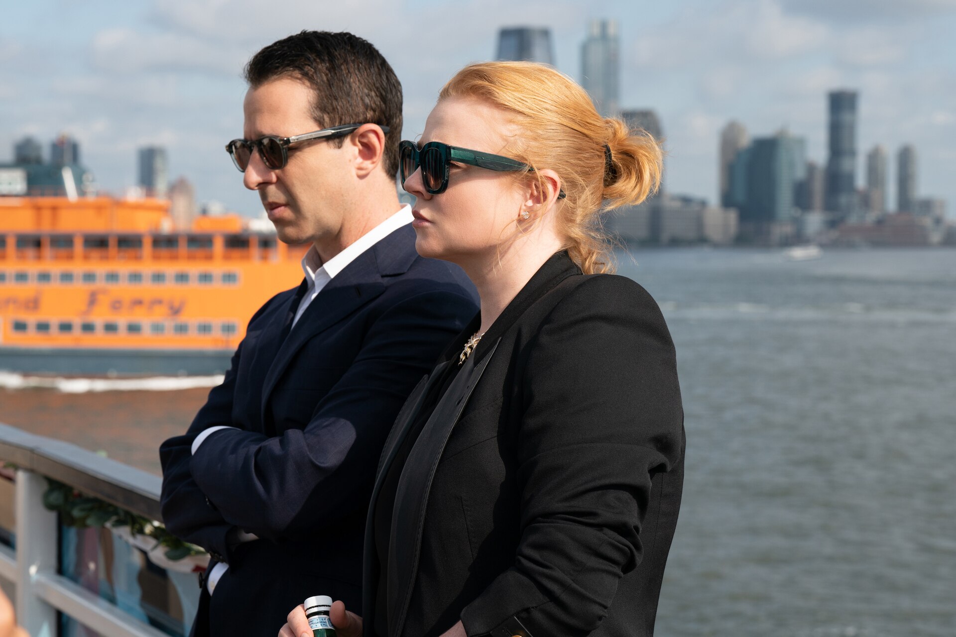 Kendall Roy and Shiv Roy, dressed in black and wearing sunglasses, sport serious expressions and stand side by side on a boat