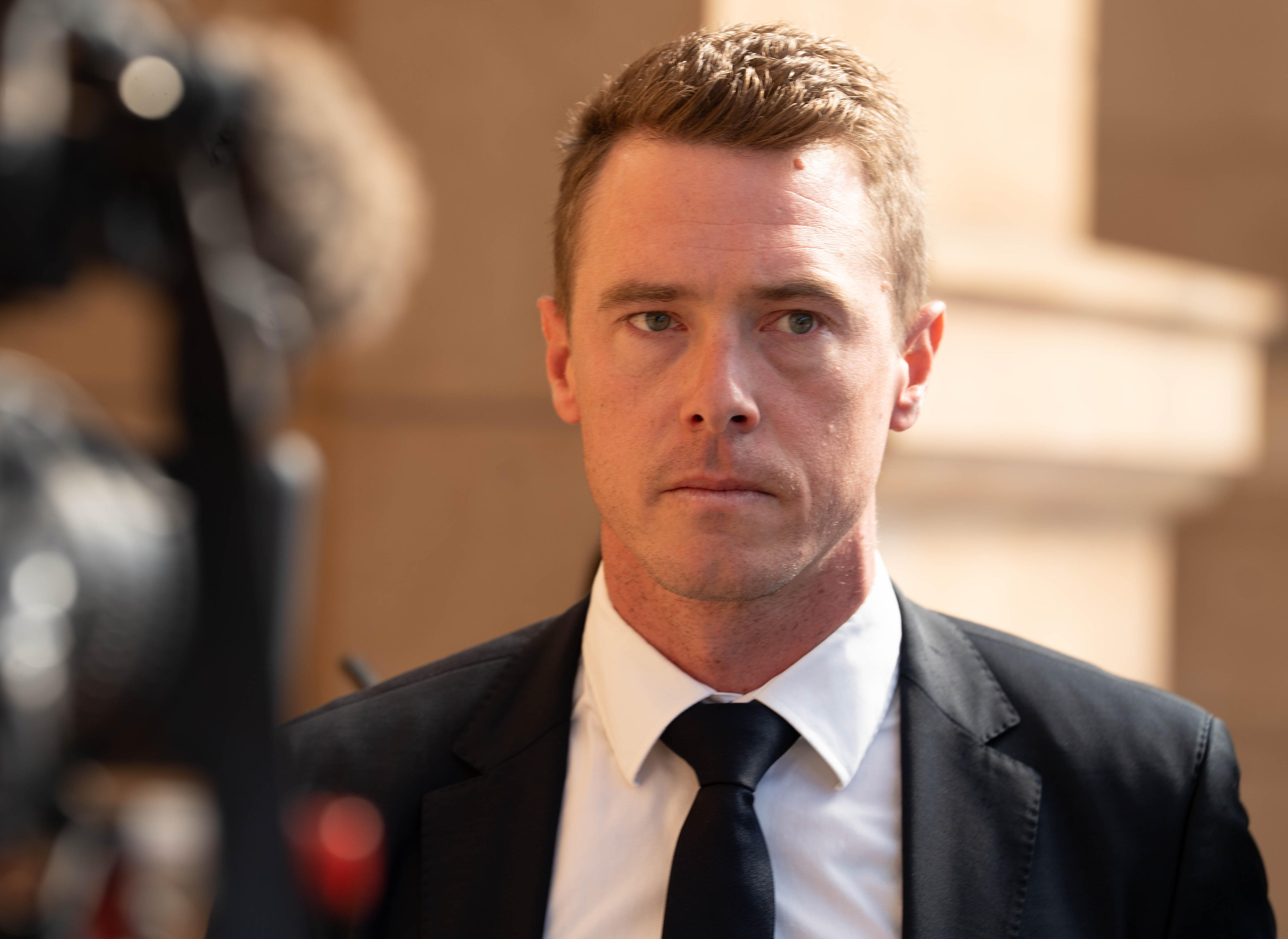 Rohan Dennis wearing formal attire walks along a sandstone wall, with a broadcast camera partially visible.