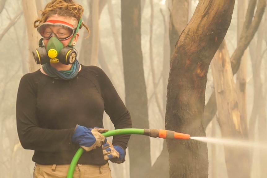 A woman holding a hose smiles at the camera whilst fighting a bush fire.