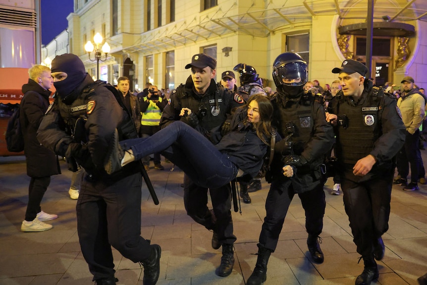 Four police officers carry a woman away from a crowd. 