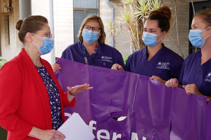 A woman with a red blazer talking to nurses in pruple scrubs holding up a purple ban