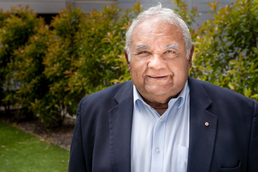 An older Aboriginal man wears a suit and smiles at the camera. There is grass and bushes behind him.