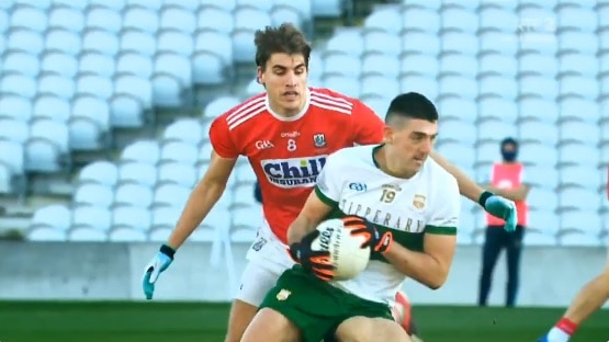 Colin O'Riordan holds a round ball in both hands wearing a white shirt with a green stripe with a man in red behind him