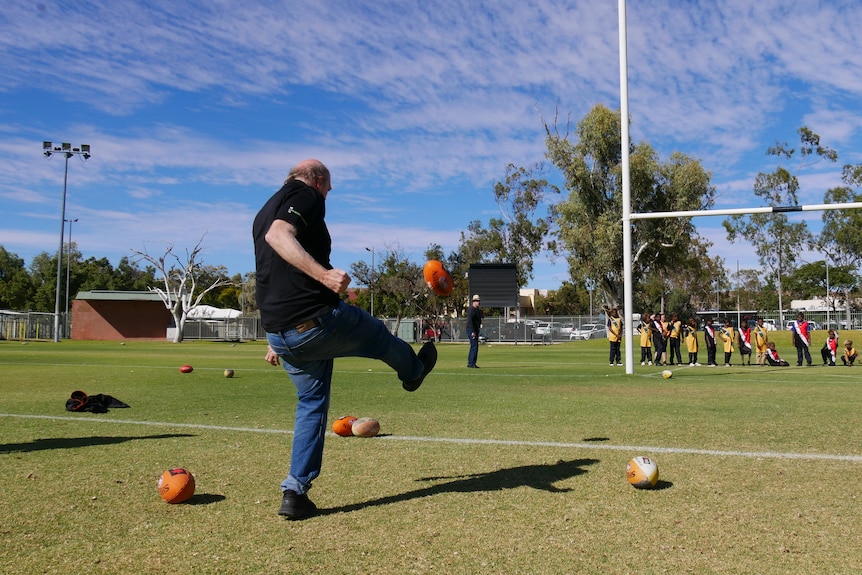 A man kicking a football towards a set of goal posts while a group of children sitting behind the posts watch on.