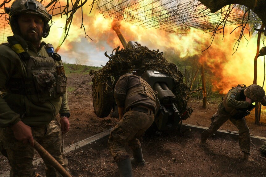 Ukrainian servicemen fire a D-30 howitzer towards Russian troops amid a cloud of flames.