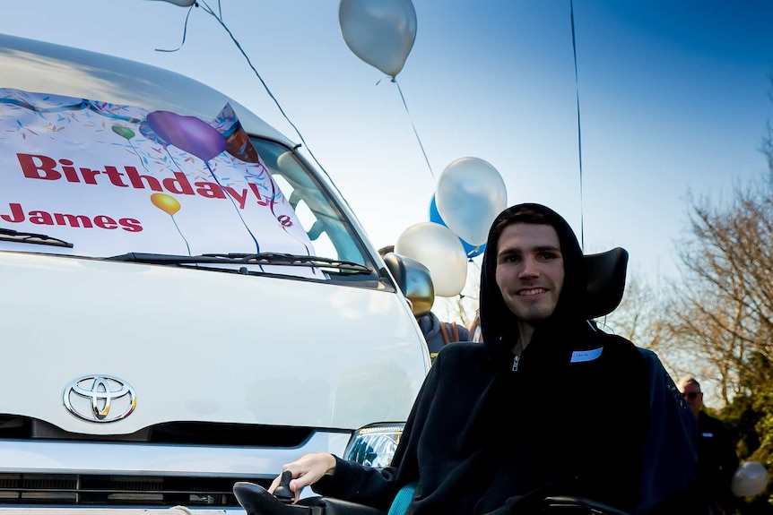 A man in a hoodie sitting a wheelchair in front of the bonnet of a van with a sign on it saying 'Birthday James'