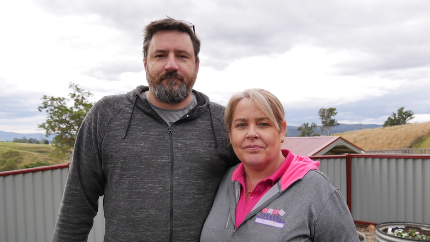 A tall middle-aged man stands in a grey jumper on the left, next to him is a shorter middle aged woman in a pink and grey jumper