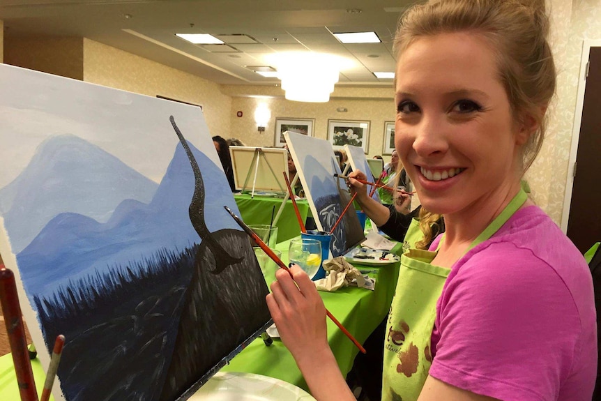 A woman smiling at the camera while holding a paintbrush to a canvas