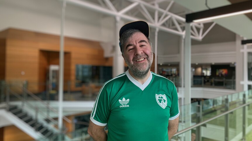 Man stands with hands behind his back, green soccer jersey and hat, smiling at camera.