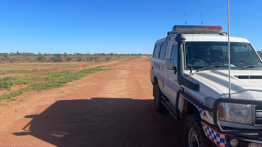 A police four-wheel drive parked on the side of a long, unsealed outback road beneath a cloudless sky.