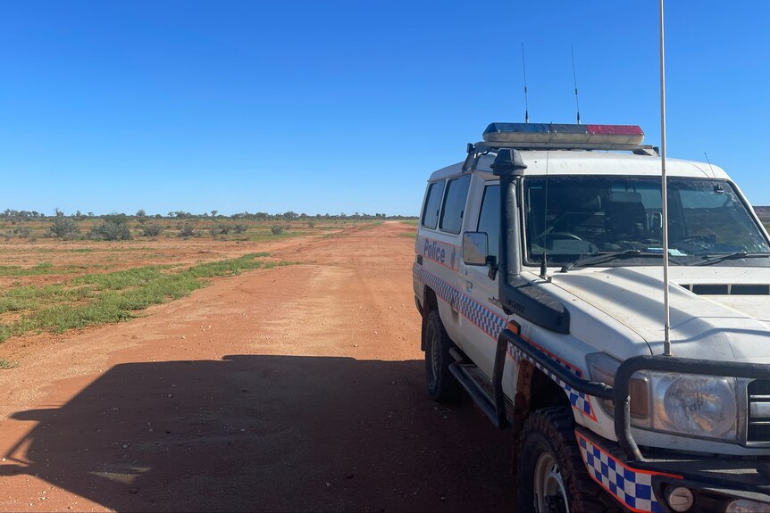 A police four-wheel drive parked on the side of a long, unsealed outback road beneath a cloudless sky.