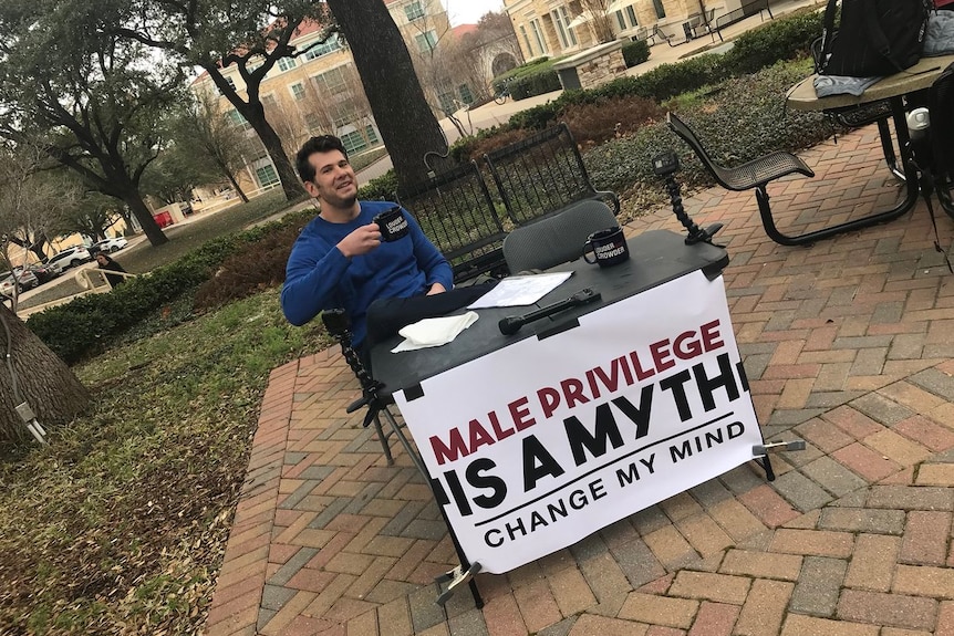 A dark-haired man sits on a campus with a sign that says "Male privilege is a myth — change my mind".