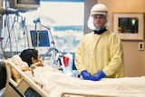 A man in a face shield and yellow hospital gown stands over a hospital bed where another man is hooked up to a ventilator