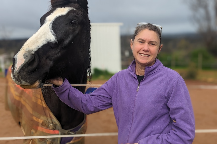 Heather Barrett pictured smiling outside and patting a black horse she is standing next to