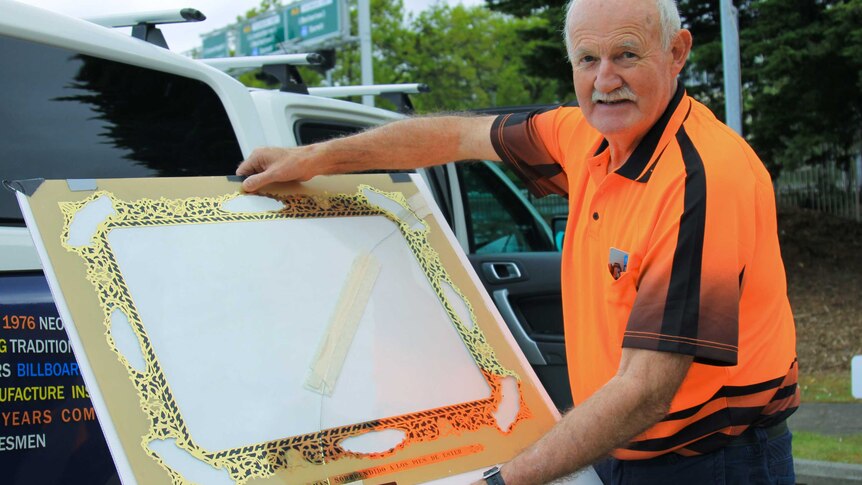 A man in a bright orange tshirt holding a large pane of glass with a black and gold design