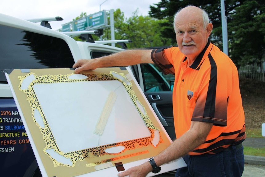 A man in a bright orange tshirt holding a large pane of glass with a black and gold design