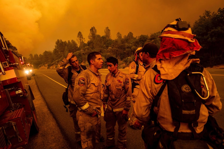 A group of firefighters stand and talk as the fire turns the sky and scene orange.