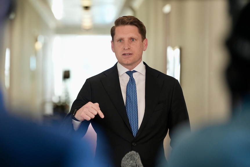 A white man with short dark hair and blue eyes pointing down as he speaks, wearing black suit and blue tie.